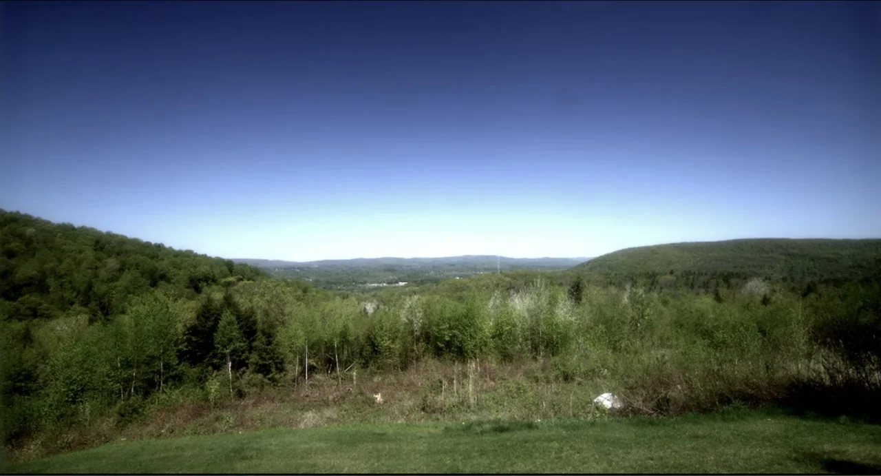 The Secret Village (2013) movie still: establishing — A vast expanse of green hills and dense forest under a clear blue sky. No human subjects or…