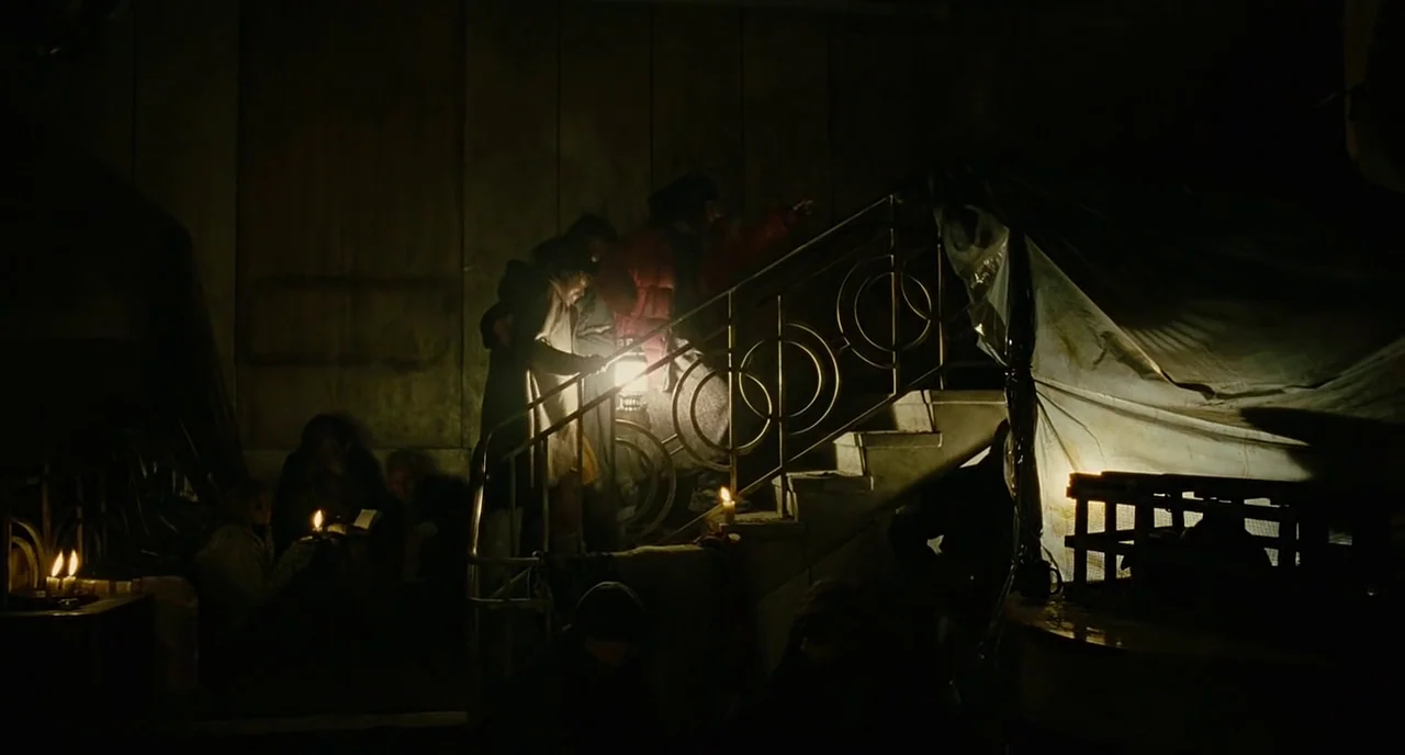 Children Of Men (2006) movie still: wide — A dimly lit indoor staircase features group members moving up with lanterns in hand. Tattered plastic…