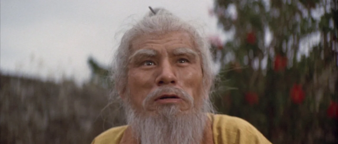 Godzilla Vs Mechagodzilla (1974) movie still: closeup — An elderly man with white facial hair is positioned outdoors near flowering foliage. He…