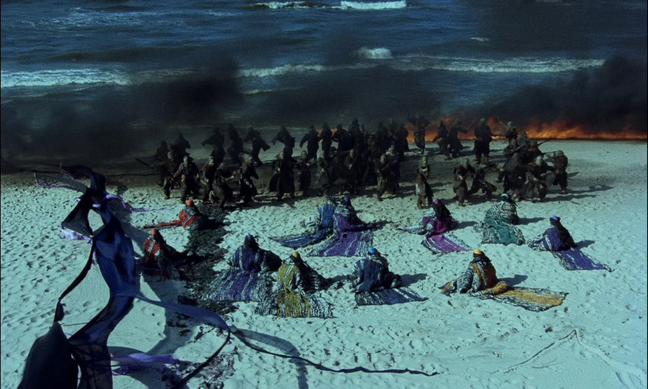 On The Silver Globe (1988) movie still: wide — A sandy beach meets the ocean with thick black smoke rising against the shoreline. Figures in…