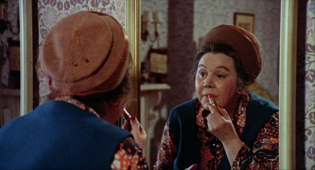 Black Christmas (1974) movie still: medium — A domestic space features wallpaper with a floral pattern and a large mirror. An older woman in a blue…