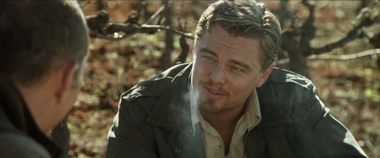 Blood Diamond (2006) movie still: medium — A man sits among dormant grape vines in a field at daytime. The subject looks at another person while…