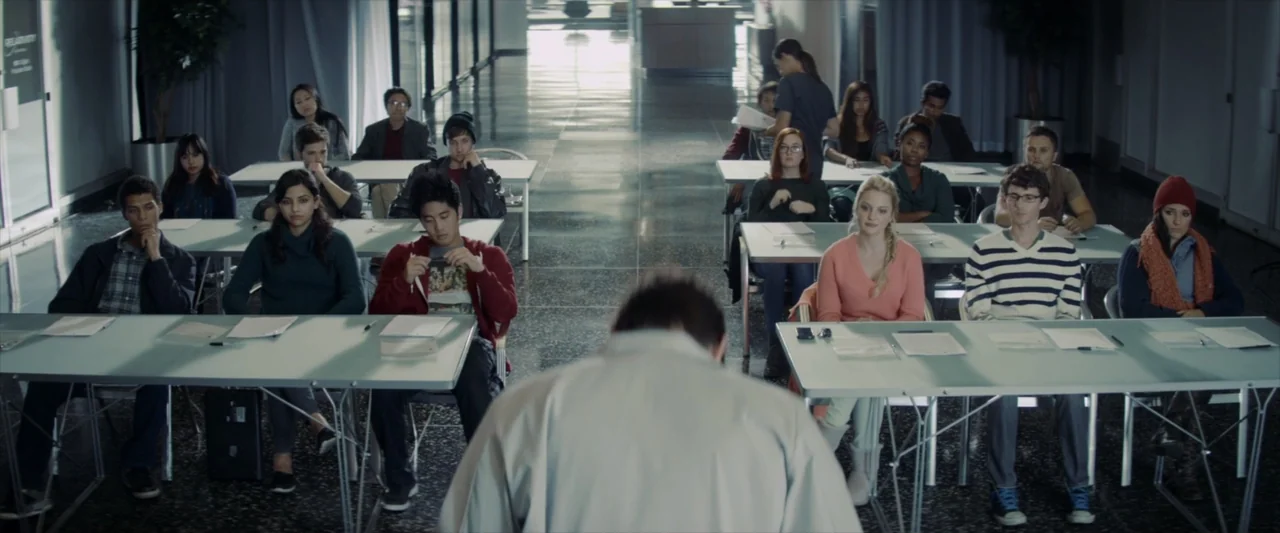 Tell Me How I Die (2016) movie still: medium — A spacious room features multiple tables arranged in rows with students sitting behind them. A person…