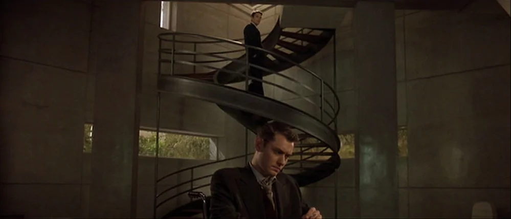 Gattaca (1997) movie still: medium — A stark, minimalist modern interior features concrete walls and a large, central spiral staircase. A man sits…