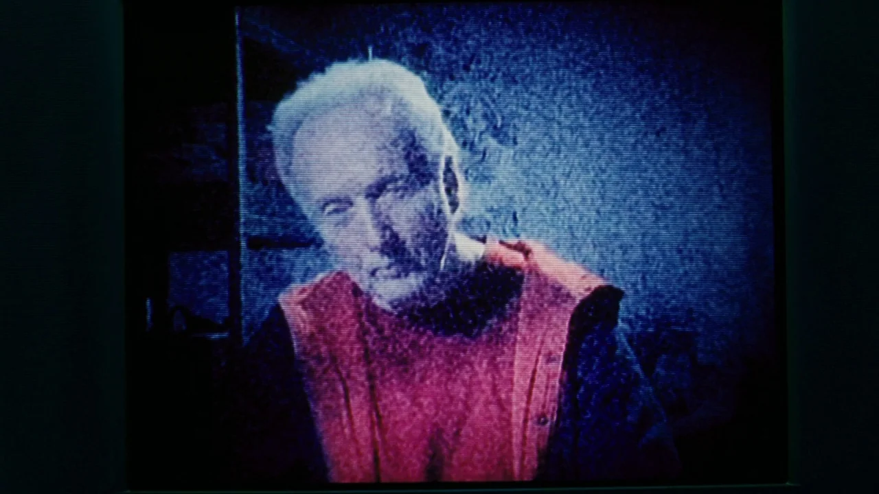 Saw VI movie still: closeup — An elderly man is displayed on a television screen with heavy scan lines. The image quality is low resolution and…