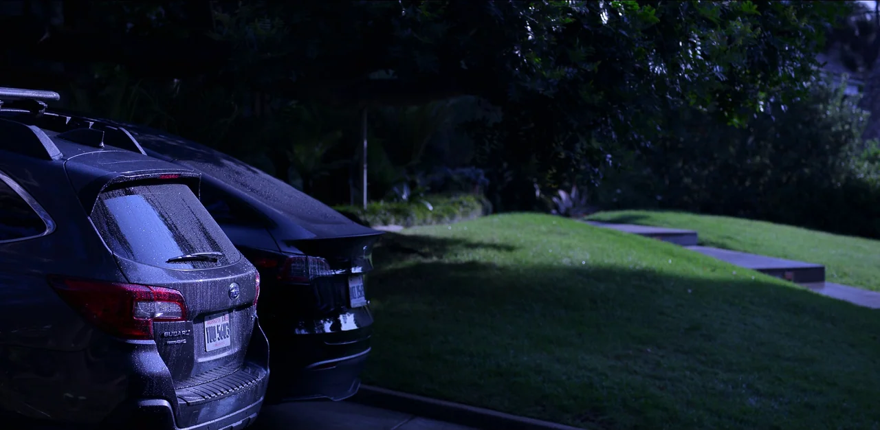 Kimi (2022) movie still: wide — A residential driveway at night features two cars parked side by side. A manicured green lawn extends from the…