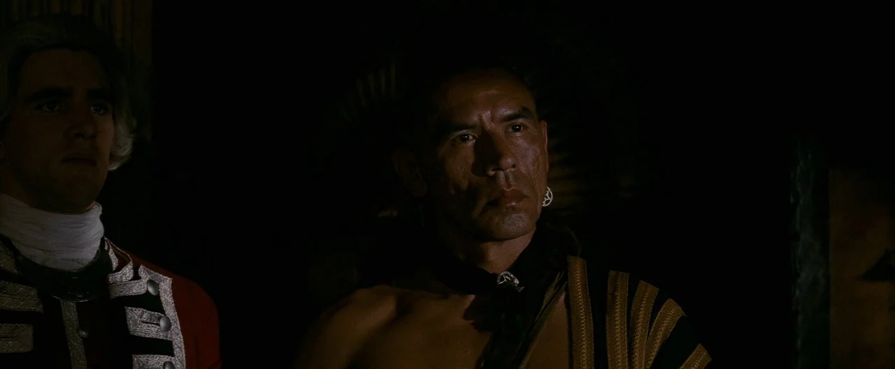 The Last of the Mohicans (1992) movie still: medium — A dimly lit interior space contains two men standing near one another. One man wears a red…