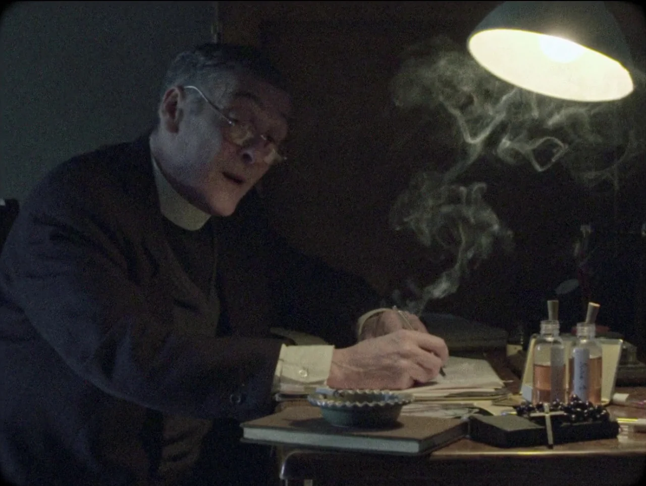 The Devil’s Doorway (2018) movie still: medium — A dark, dimly lit study features a wooden desk cluttered with papers and books. An elderly man in…