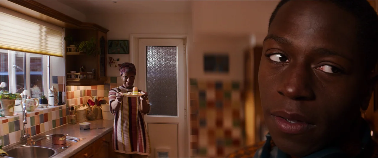Rye Lane (2023) movie still: medium — A home kitchen with multicolor wall tiles and a window overlooking an exterior view. A woman standing at the…