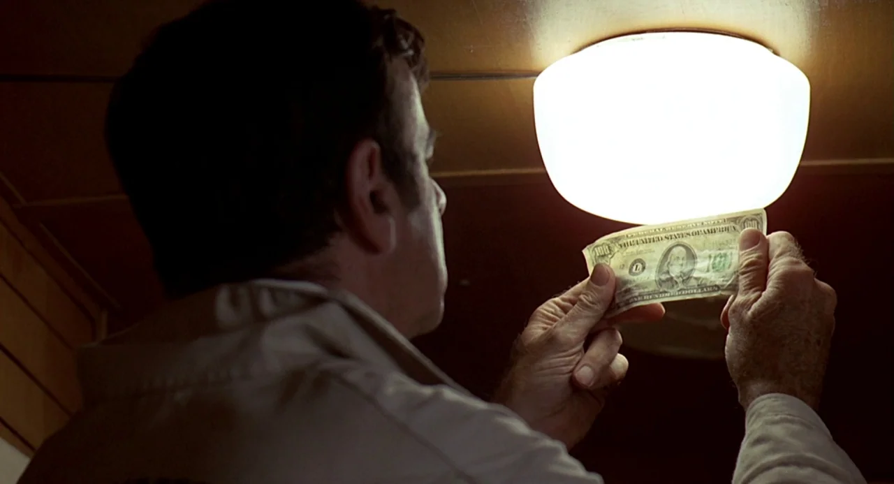 Charley Varrick (1973) movie still: medium — A man in a tan jacket holds a banknote up to a bright ceiling lamp. The scene is illuminated by the…