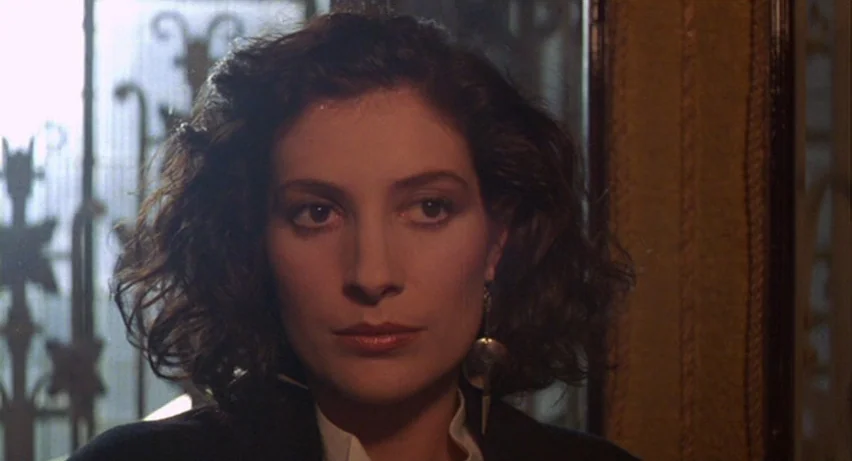 Matador (1986) movie still: closeup — An interior space with blurred ornate wrought iron detail visible through a window. A woman looks forward…
