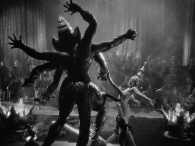Mata Hari (1931) movie still: fullbody — An opulent ballroom holds a large multi-armed deity statue in the foreground. A performer dances to the…