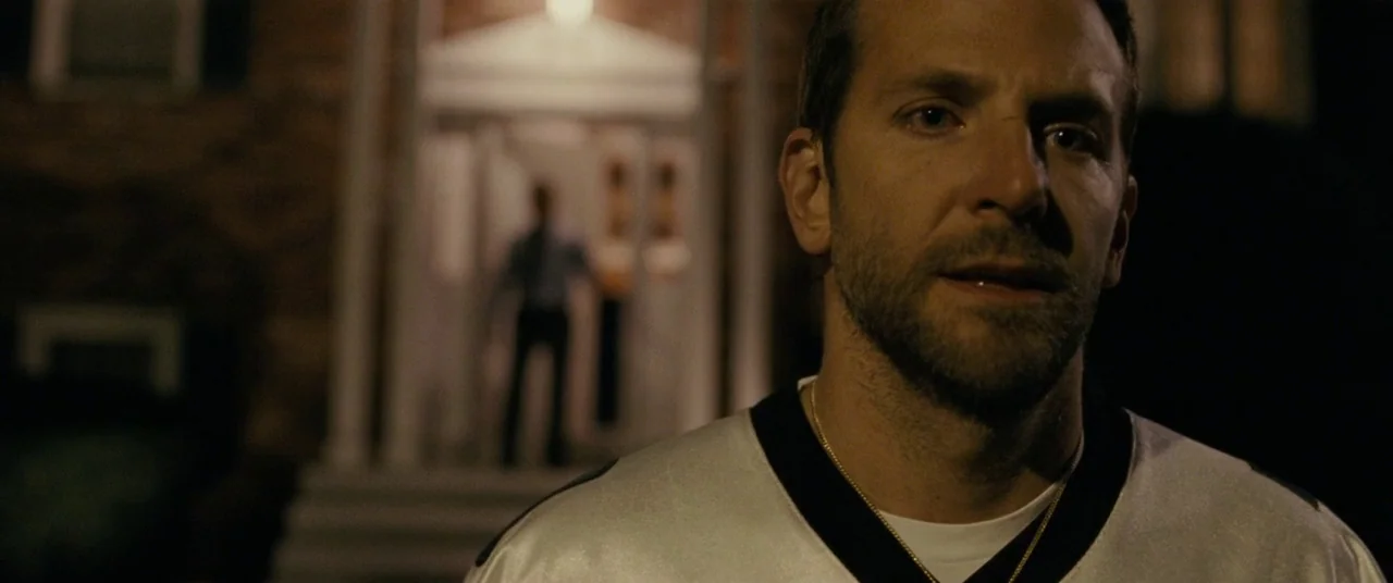 Silver Linings Playbook (2012) movie still: closeup — A man in an athletic jersey stands in the dark foreground at night. Another man is visible in…