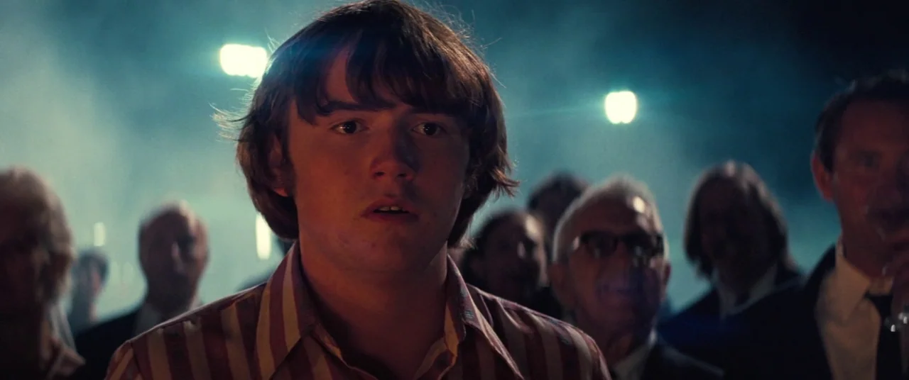 Licorice Pizza (2021) movie still: closeup — A teenage boy stands in a crowd during a dimly lit night event outdoors. He faces the camera directly…
