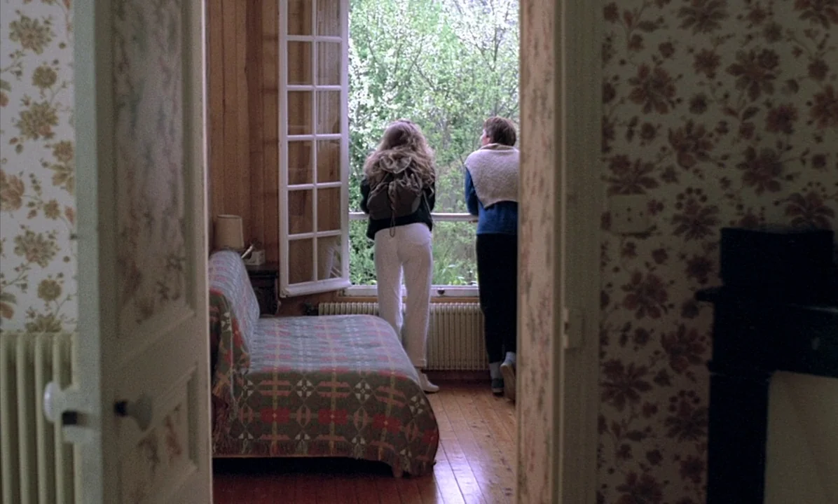 A Tale of Springtime (1990) movie still: wide — A bedroom features floral wallpaper, a patterned daybed, and hardwood floors. Two people stand at an…
