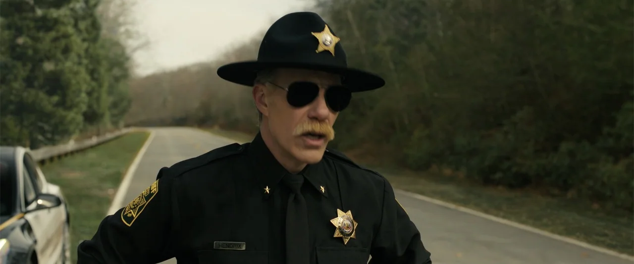 Companion (2025) movie still: medium — A quiet rural road bordered by trees features a law enforcement officer standing nearby. The officer wears a…