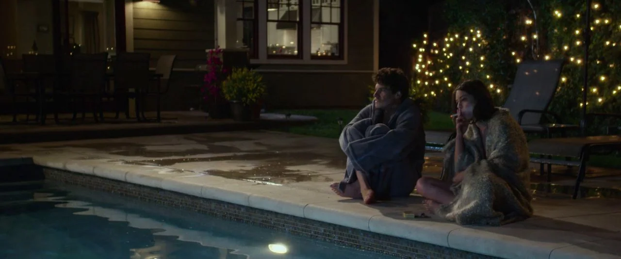 Night Owls (2015) movie still: wide — A dark outdoor pool deck features a house in the background with lit windows. A man and woman sit on the tiled…