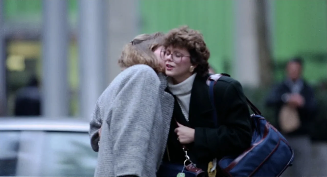 Baby Boom (1987) movie still: medium — A city street background features a blurred green facade and a car hood. Two women wearing coats embrace…