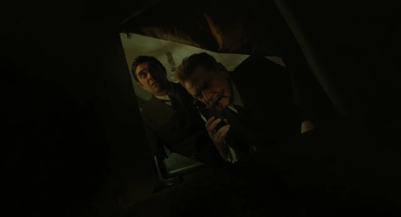 A Haunting In Venice (2023) movie still: medium — Two men in suits stand in a dimly lit, enclosed area speaking on a telephone. The composition is…