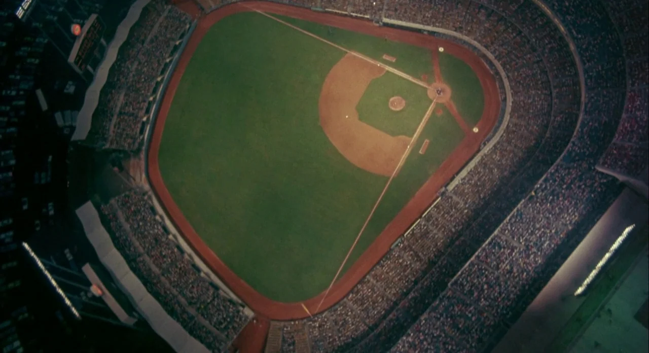 Koyaanasquatsi (1982) movie still: establishing — An aerial overhead view shows a professional baseball stadium with bright artificial lights. The…