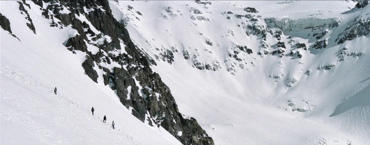 Society of the Snow (2023) movie still: wide — A vast, snow-covered mountainside features a steep, jagged rock face to the left. Four small figures…