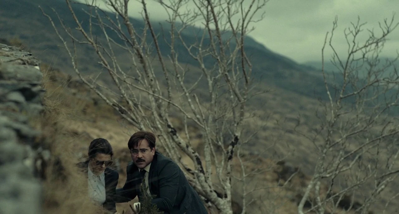 The Lobster (2015) movie still: medium — A bleak, windswept hillside features dry grass and bare, leafless trees against distant mountains. A man in…
