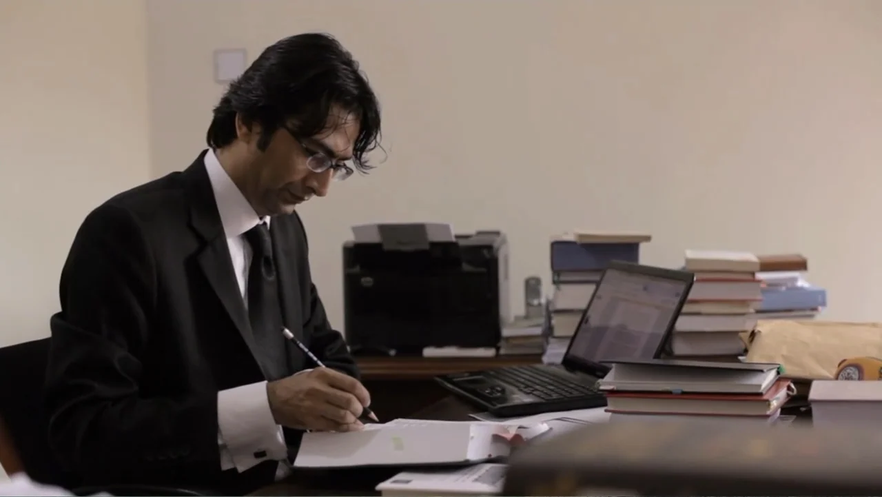 A Girl in the River: The Price of Forgiveness (2015) movie still: medium — A man works at a desk cluttered with stacks of books and papers. The…
