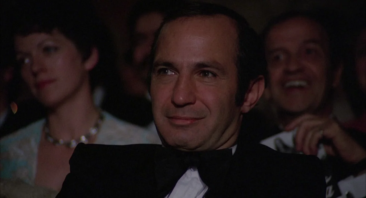 Opening Night (1977) movie still: closeup — A man in a tuxedo sits in a dark, crowded theater during an event. He looks toward the front while…