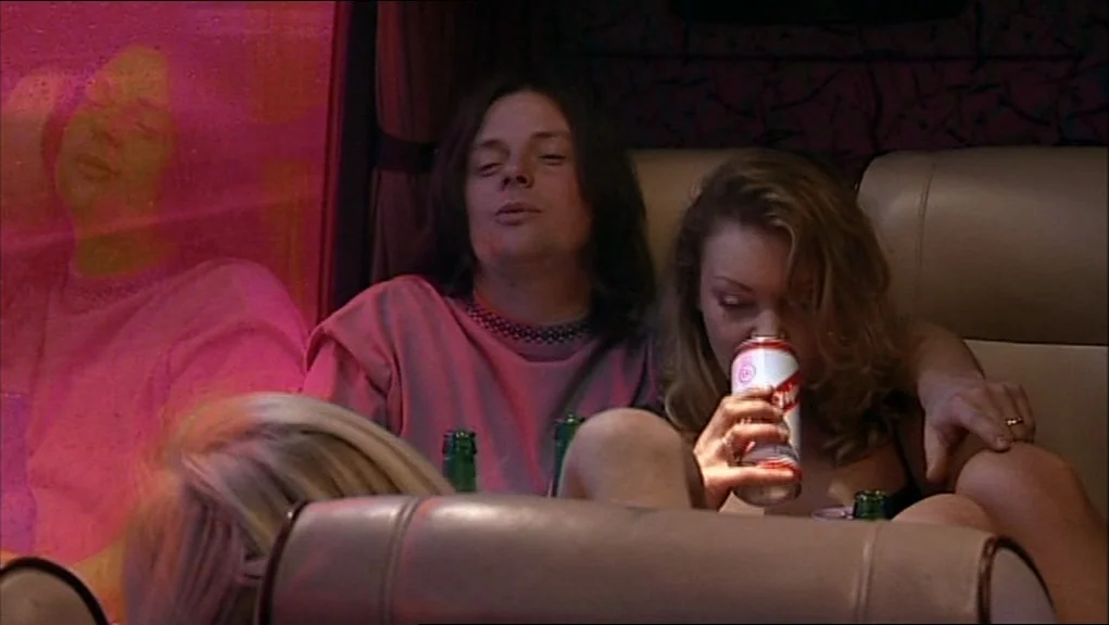 24 Hour Party People movie still: medium — The interior of a bus features dark leather seats and reflective tinted windows. A man and a woman share…