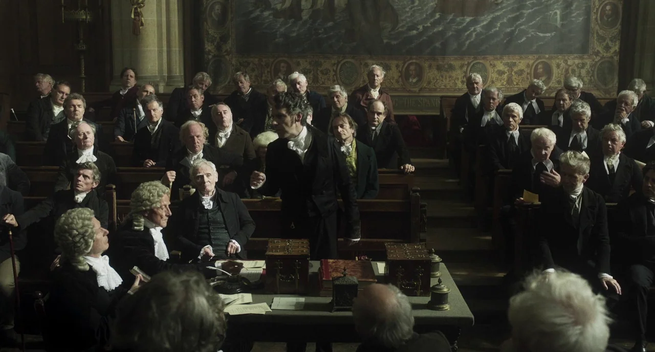 Peterloo (2018) movie still: wide — A historic courtroom with wooden benches and a large tapestry hanging on the back wall. Men in period tailcoats…