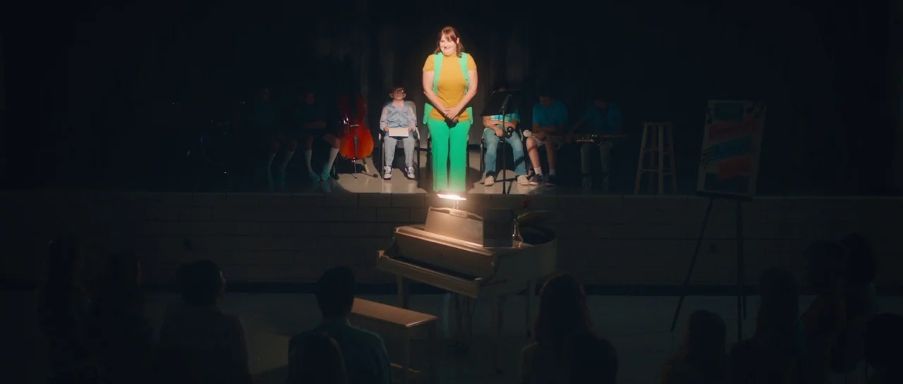 Greener Grass (2019) movie still: wide — A dimly lit school auditorium features a stage with students and a teacher present. The teacher stands in a…
