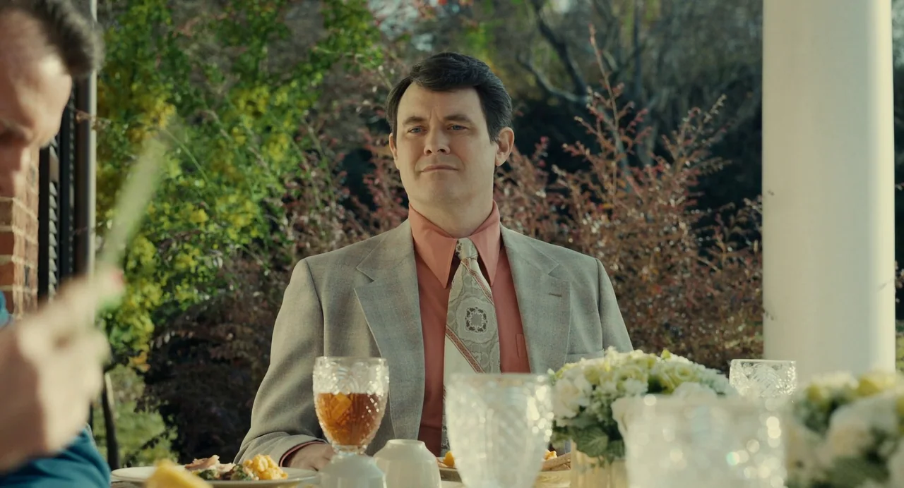 The Eyes of Tammy Faye (2021) movie still: medium — A man sits at a patio table during the day with lush greenery behind him. He wears a formal suit…