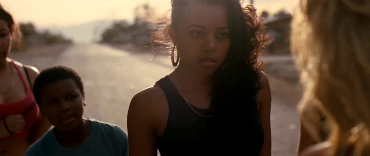 Little Birds (2011) movie still: medium — A residential street in the late afternoon sun with a group of people gathered. A woman in a black tank…