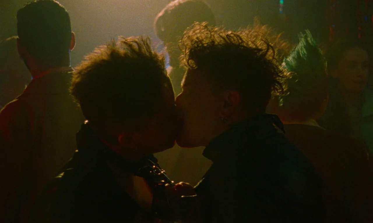 Blue Jean (2022) movie still: medium — A crowded, dimly lit nightclub features people gathered under hazy, golden atmospheric lights. Two…