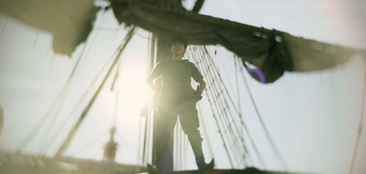 Peter Pan & Wendy (2023) movie still: fullbody — A person stands on the wooden deck of a tall ship with tattered sails overhead. The sun shines…