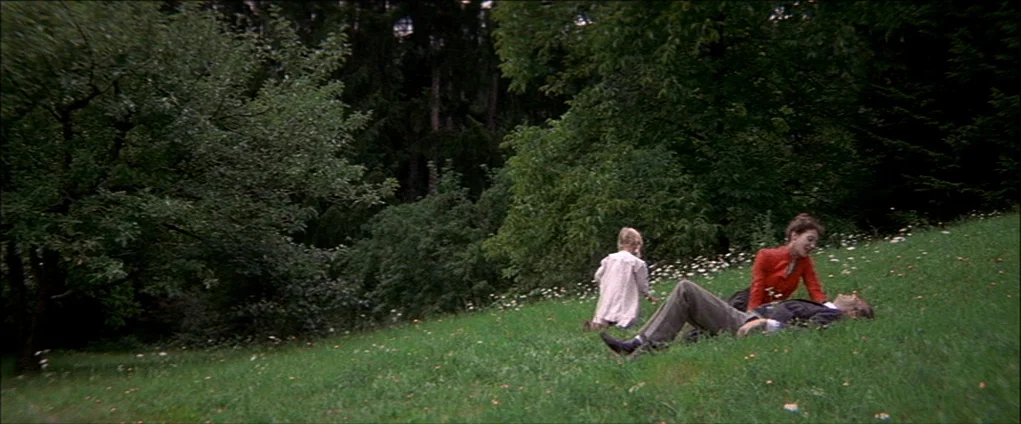 Death In Venice (1971) movie still: wide — A grassy hill slopes upward toward a dense forest of dark trees. A family group rests on the hillside…