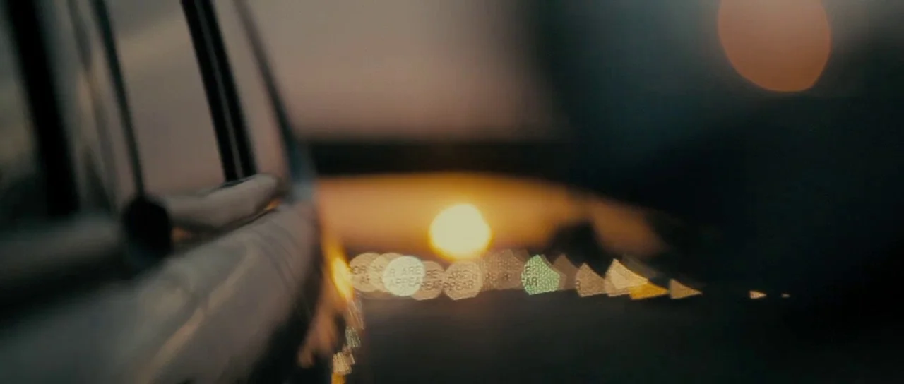 The Imposter (2012) movie still: closeup — A close view of a car door frame during the late golden hour. Out of focus street lights create a line of…