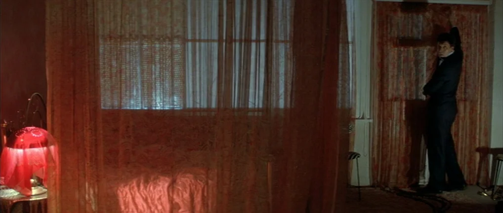 The Crying Game (1992) movie still: wide — A dark, cramped space features heavy, patterned curtains draped over a window. A man in a suit stands by…