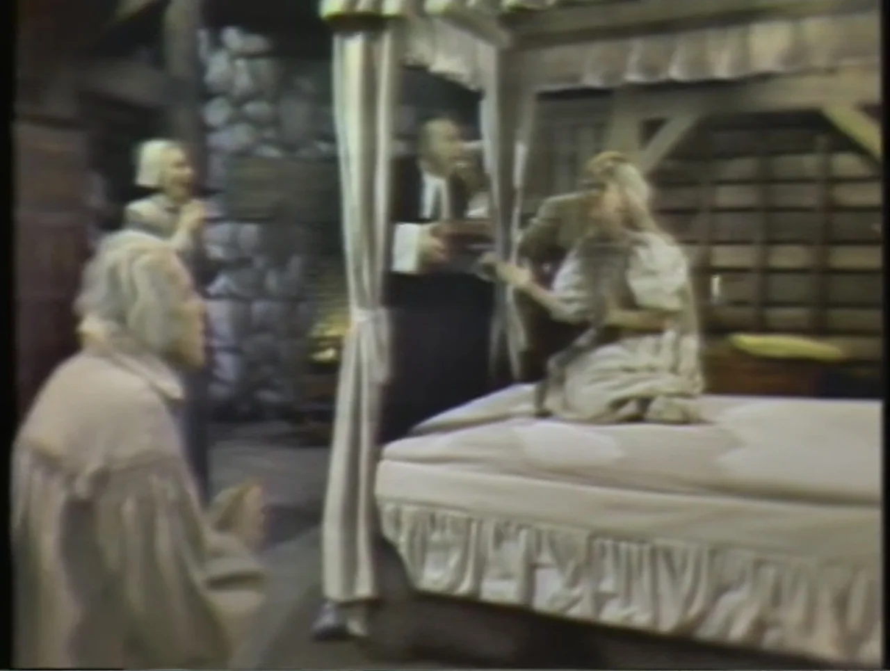 The Crucible (1996) movie still: fullbody — A stone-walled interior contains a large wooden canopy bed draped in fabric. A young woman kneels upon…