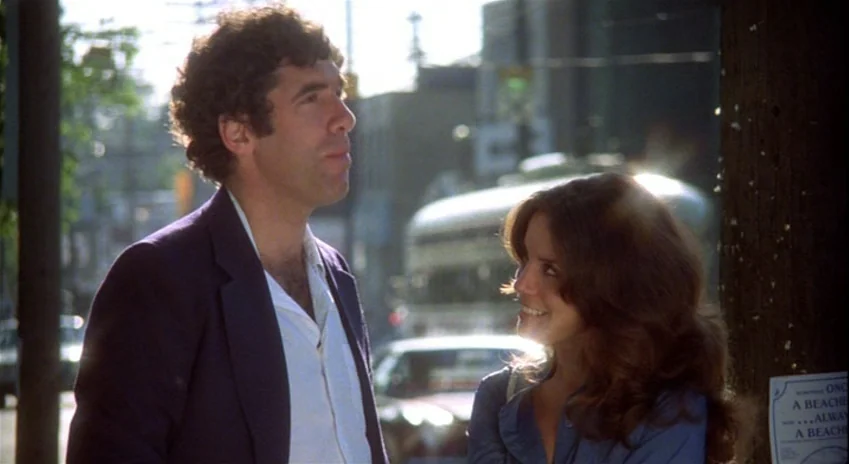 The Silent Partner (1978) movie still: medium — An urban sidewalk backdrop with a blurred street and passing cars behind the couple. A man in a…