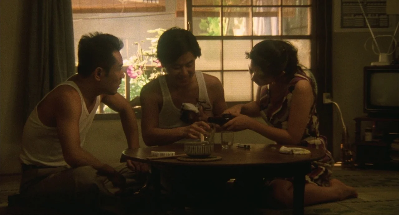 The Discarnates (1988) movie still: medium — A low-lit apartment with a window and a round wooden low table. Two men and a woman sit around the…