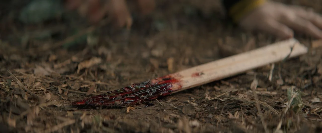 Becky (2020) movie still: closeup — A wooden stake rests on a dirt and leaf-covered forest floor. The tip of the wood is heavily coated in dark red…