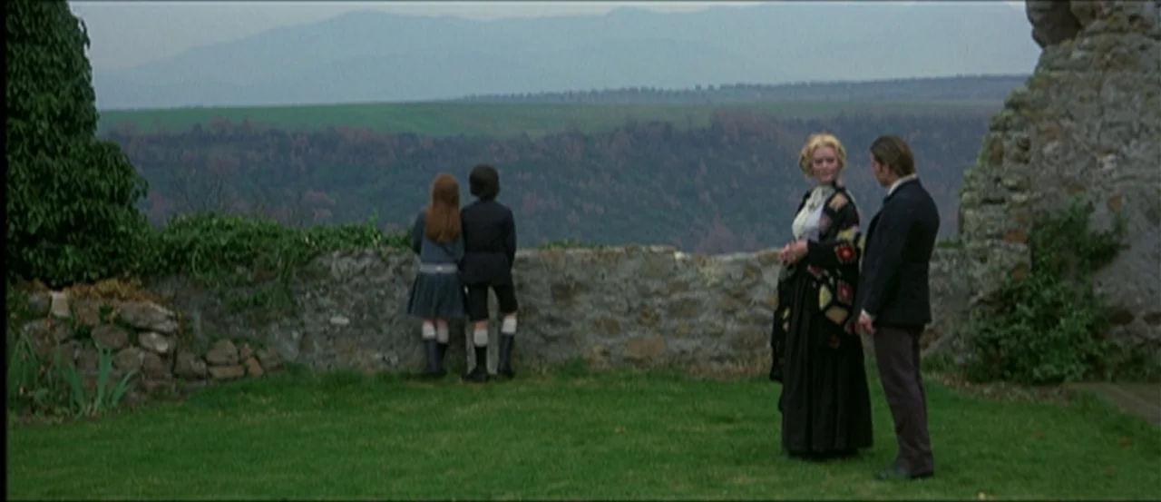 Flesh For Frankenstein (1973) movie still: wide — A stone wall separates a grassy lawn from a wide view of distant mountains. Two adults stand on…