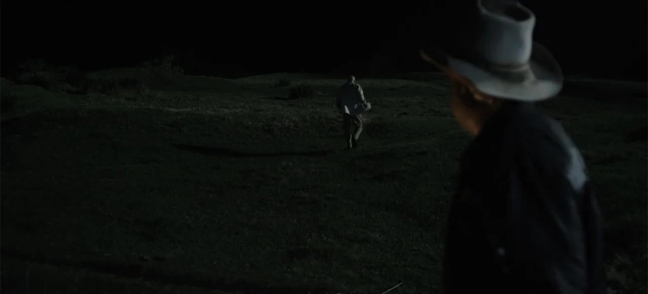 Boar (2018) movie still: wide — A dark, rolling open field under a pitch-black night sky. A man walks away into the distance while another man…