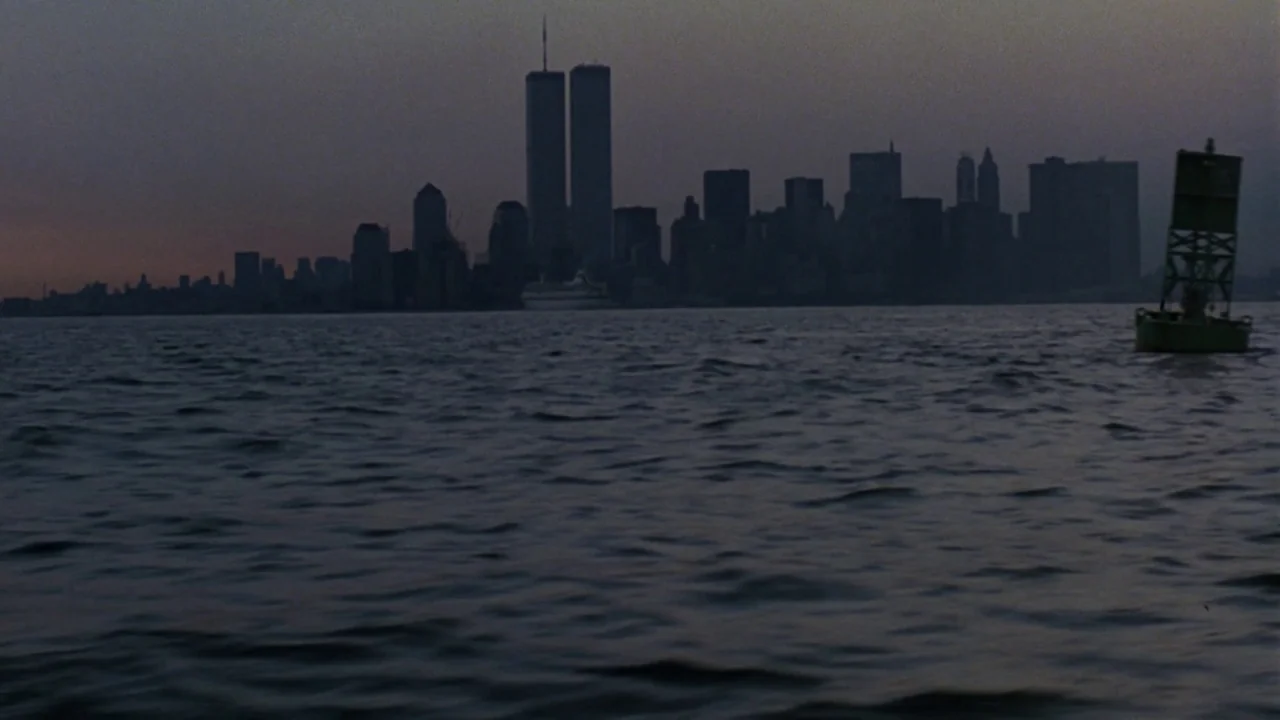 Something Wild (1986) movie still: establishing — A harbor view looks toward a dark Manhattan skyline at twilight. A green navigational buoy floats…