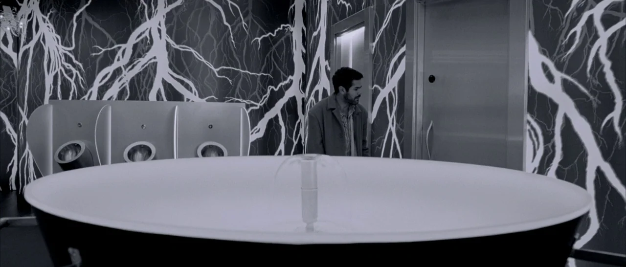 Angel-A (2005) movie still: medium — A modern public restroom features black walls with white lightning-bolt patterns. A man stands beside a…