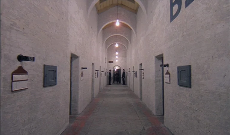 A Clockwork Orange (1971) movie still: establishing — A long symmetrical hallway lined with identical doorways leads to a central gated entrance.…