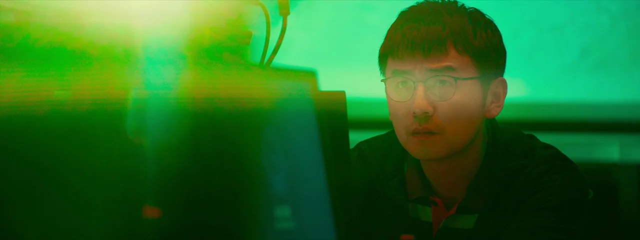 Under The Light (2023) movie still: medium — A workspace illuminated by the intense green light of a computer monitor at night. A man wearing…