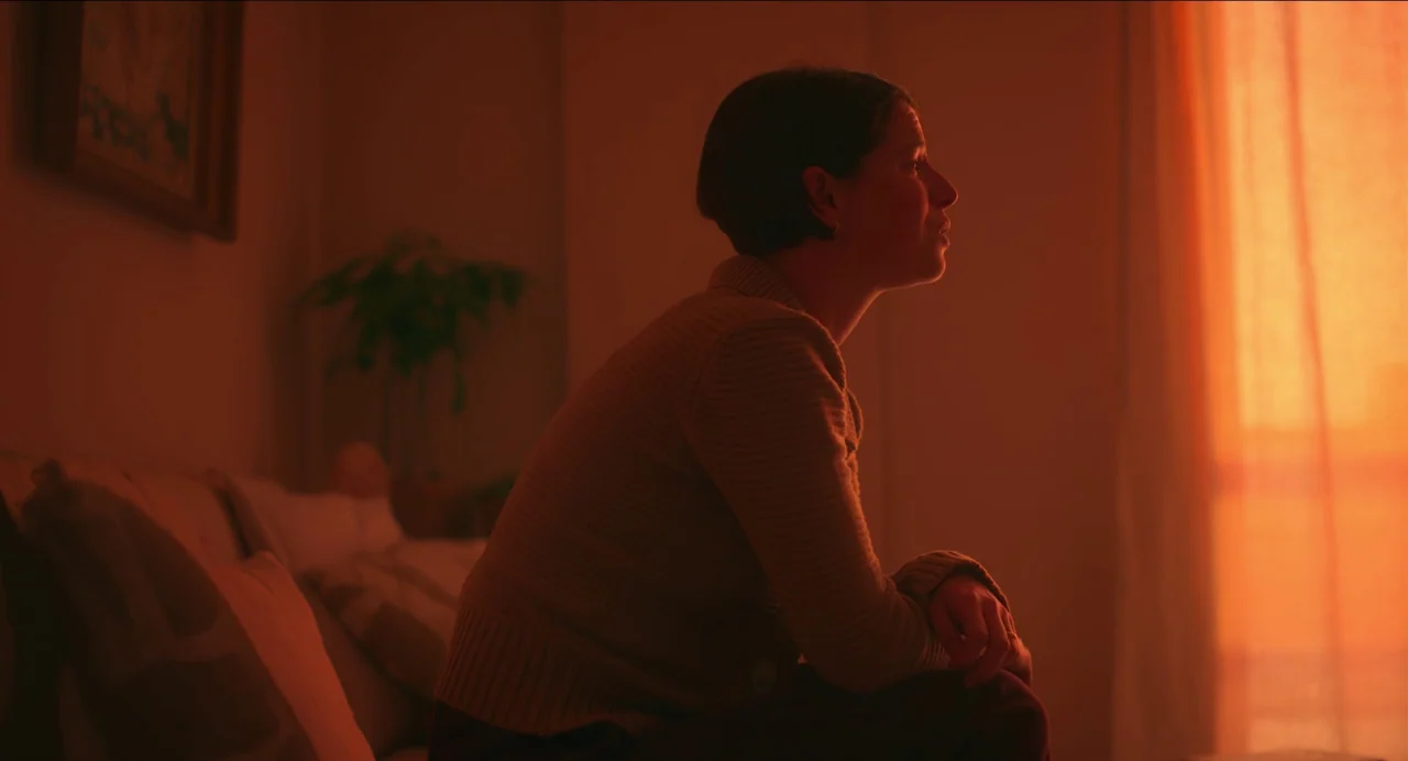 Men (2022) movie still: medium — A dimly lit room with a soft orange glow emanating from a nearby window. A woman with short hair sits on a couch,…