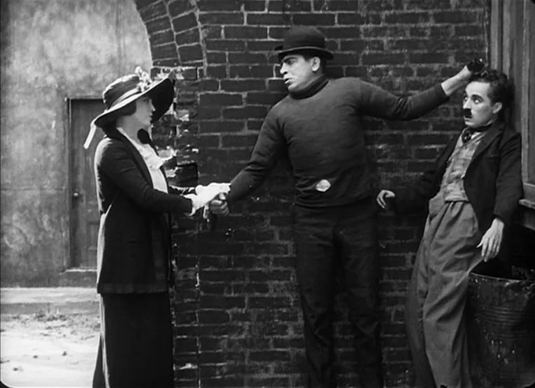 The Kid (1921) movie still: medium — A woman stands in a paved outdoor area before a brick wall. A man in a sweater holds her hand while the other…